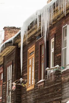 Sharp transparent icicles hang on the edge of the roof. Against the background of the wooden wall of the old house. Large cascades, even beautiful rows. Cloudy winter day, soft light.