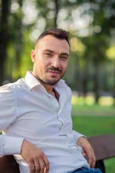 A happy business man in a white shirt is resting while sitting on a bench. Young man in city park, warm sunny summer day. Warm soft light, close-up.