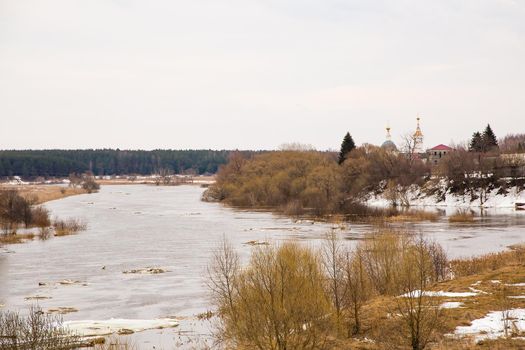 Pieces of white ice floes float down the river. Spring, snow melts, dry grass all around, floods begin and the river overflows. Day, cloudy weather, soft warm light.