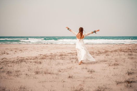 Model in boho style in a white long dress and silver jewelry on the beach. Her hair is braided, and there are many bracelets on her arms