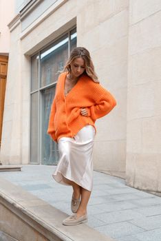 Portrait of fashionable blond women in orange sweater, beige dress and stylish suede loafer shoes posing in the street