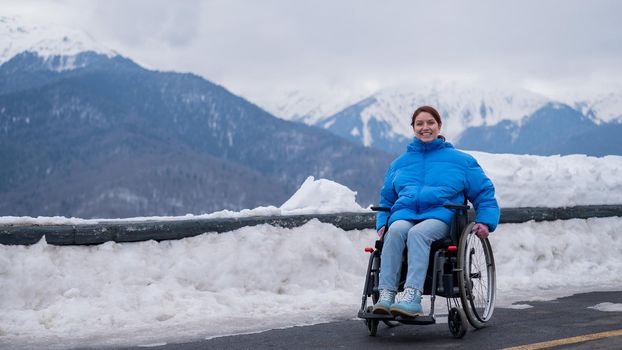 A happy woman dressed in a blue coat sits in a wheelchair on a point view and looking at the snow-capped mountains