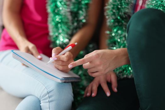 Close-up of women writing wish list for christmas, empty page to list desires for gifts. Creative atmosphere before new year, presents under christmas tree. Holiday concept. Blurred background