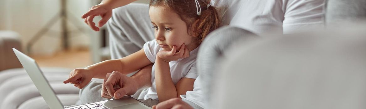 Little daughter is sitting on father laps while spending time on couch with parents and brother at home