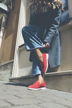 Woman in a red stylish suede loafer shoes, blue jeanse, white shirt and trendy checkered coat. Pair of trendy female loafers shoes, outdoors