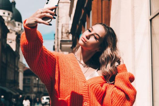 Charming happy woman student communicates by phone. Portrait of fashionable women in orange sweater and beige dress making selfie on the street