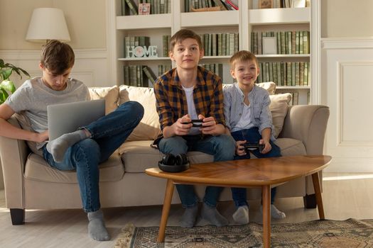 Three brother boys are sitting at home on a sofa in the sun playing computer games on a laptop and video games