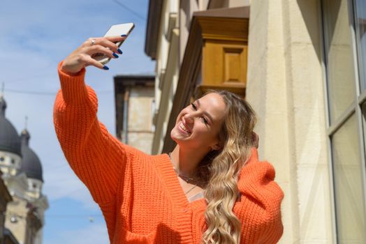 Charming happy woman student communicates by phone. Portrait of fashionable women in orange sweater and beige dress making selfie on the street