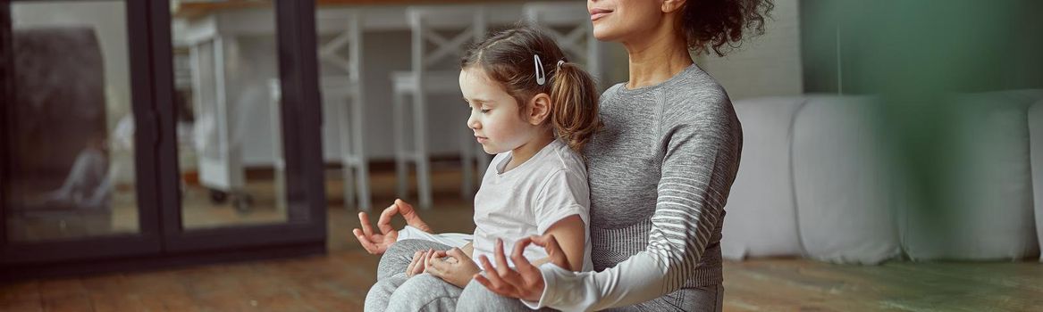 Relaxed woman is sitting with little girl in lotus position and doing meditation at home