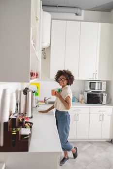 Full-length view of African American lady standing on office kitchen while holding cup with hot drink