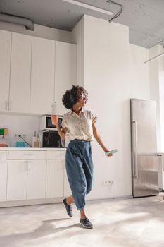 Full-length photo of smiling African American lady using headphones and dancing on the kitchen while holding smartphone