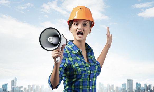 Expressive woman in safety helmet shouting into megaphone. Portrait of young emotional construction worker with wide open mouth on cityscape background. News announcement and advertisement.