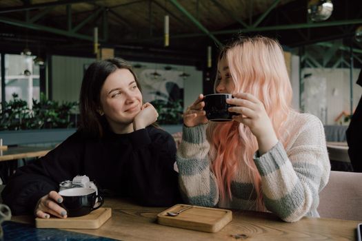 Interracial lesbian couple each other with shy smile, holding hands during lunch at restaurant. Happy redhead woman confessing love to her stylish Asian American girlfriend with pink hair hairstyle