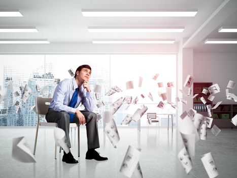 young businessman sits on an office chair. thoughtful and looking up. documents fly around