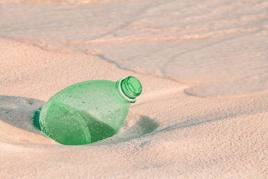 Close up of a green plastic water bottle buried in the sandy beach