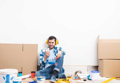 Happy boy in protective headphones sitting on floor. Home remodeling after moving. Construction tools and materials for building. Young man in casual clothes holding smartphone and paint roller