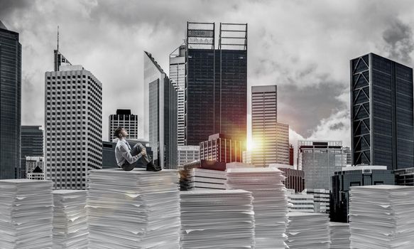 Thoughtful businessman sitting on pile of documents and looking away with cityscape and sunlight on background. Mixed media.