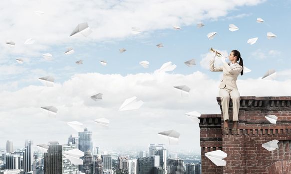 Beautiful young woman with trumpet on roof. Brave girl in white business suit with music brass instrument sitting on edge of roof. Flying paper planes in blue sky. Female musician playing music