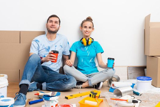 Happy smiling couple relaxing on floor with coffee after moving in new house. House remodeling and interior renovation concept. Young man and woman together sitting on floor among cardboard boxes.