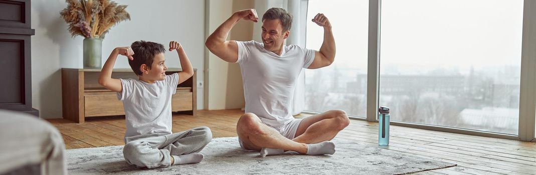 Happy man is sitting with boy on mat and showing biceps after doing workout together