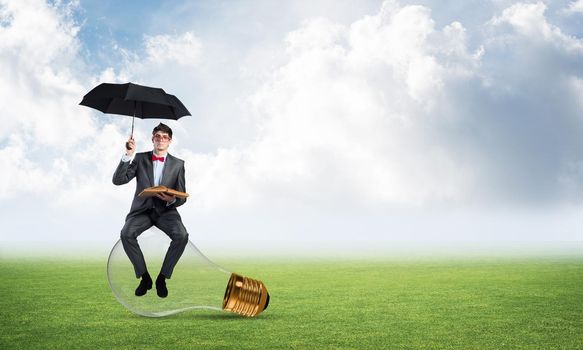 young businessman with an umbrella and a book, sitting on a light bulb. concept of finding new ideas in business