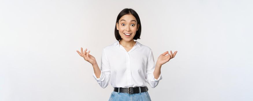 Excited young asian woman, looking happy in disbelief, hear surprising news, amazing info, standing over white background.