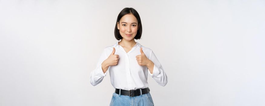 Image of beautiful adult asian woman showing thumbs up, wearing formal office, university clothing, recommending company, standing over white background.