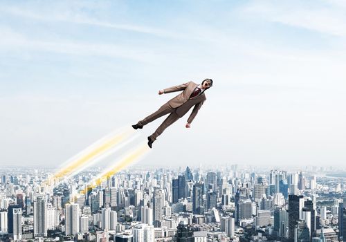 Businessman in suit and aviator hat flying in blue sky as superhero. Business person as superman launching upwards with jet flame above modern downtown. Successful business startup. Career growth