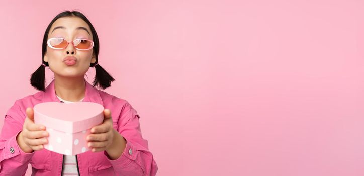 Cute asian girl giving you gift in heart shaped box, kissing and smiling, concept of holiday and celebration, standing over pink background.