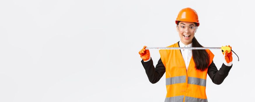 Excited and upbeat asian female construction engineer measuring layout, holding tape measure and smiling, ready for work at build something, standing over white background in safety helmet.