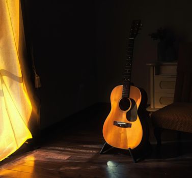 Acoustic Spanish guitar on a stand in the moody shadows of a dark room with bright light coming in from behind a curtain