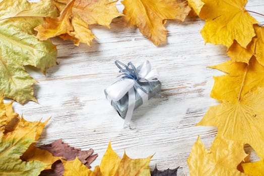 Bright autumn composition with gift box and yellow maple leaves. Holiday present decorated white ribbon bow lies on vintage wooden desk. Happy thanksgiving congratulation. Autumn sale advertising.