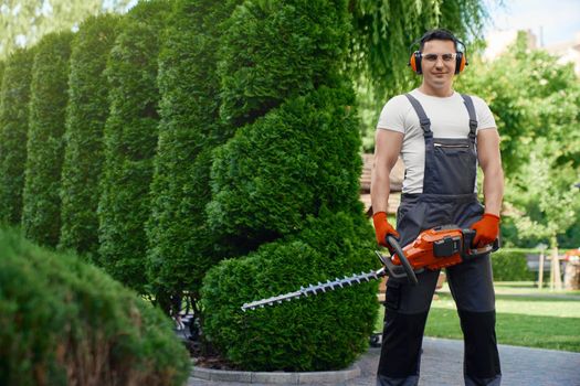 Competent caucasian gardener smiling and looking at camera while standing on back yard and holding hand electric trimmer. Man using gardening tool for cutting hedge.
