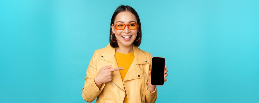 Image of young stylish asian female model in trendy sunglasses, showing mobile phone screen, application on smartphone interface, standing over blue background.