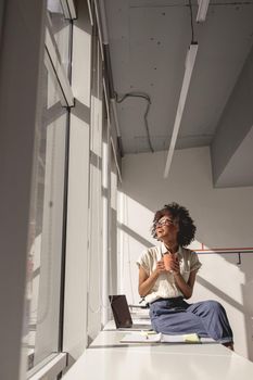 Happy African American woman enjoying sunny day in the office while holding cup of coffee