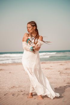 Model in boho style in a white long dress and silver jewelry on the beach. Her hair is braided, and there are many bracelets on her arms