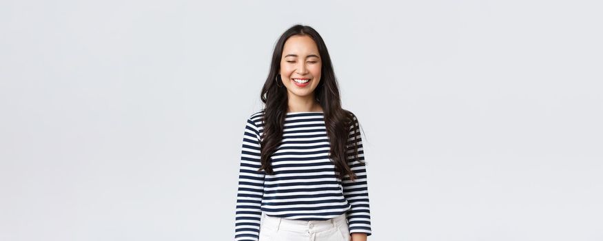 Lifestyle, people emotions and casual concept. Carefree, happy pretty asian woman in striped shirt close eyes and laughing sincere, having fun, funny moment, standing white background.