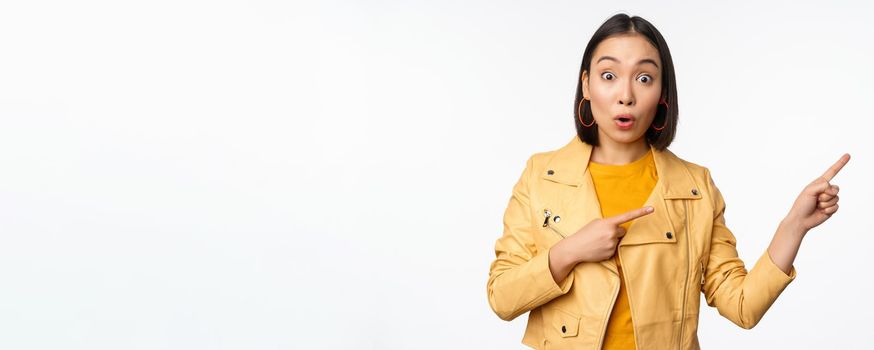 Enthusiastic asian girl pointing fingers right, showing advertisement and smiling, inviting to the store, standing over white background.