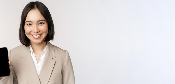 Portrait of corporate asian woman showing smartphone app interface, mobile phone screen, standing over white background.