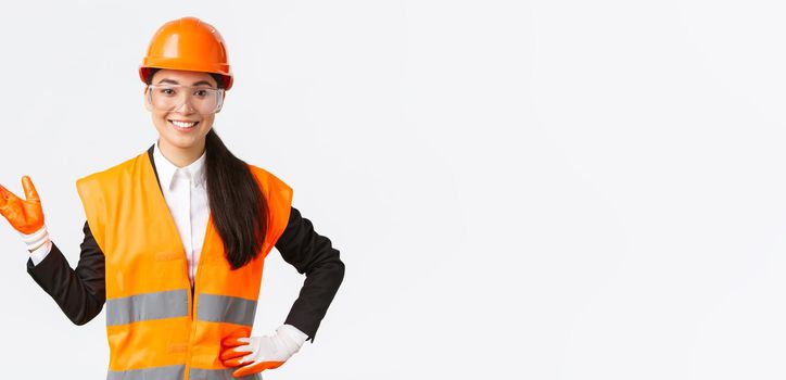Smiling cheerful asian female construction manager introduce her interprise for investors, wearing safety glasses, helmet and gloves pointing hand left and looking proud, white background.