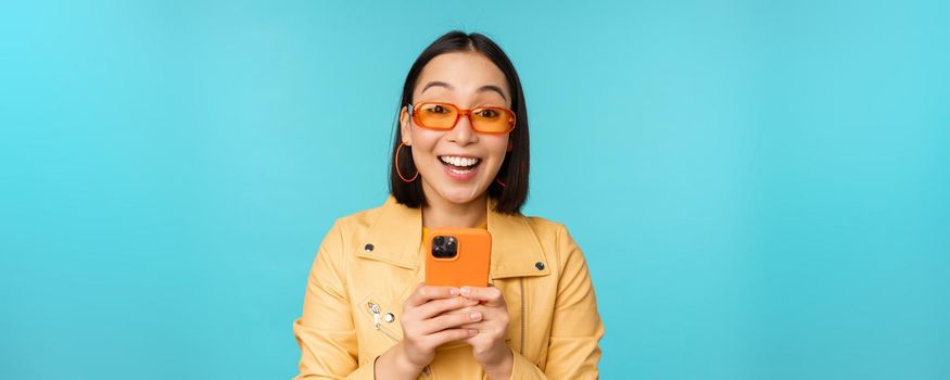 Portrait of enthusiastic asian woman in sunglasses, using mobile phone, smiling and laughing, looking happy, holding smartphone, standing over blue background.