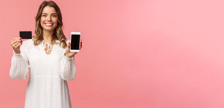 Portrait of satisfied cute blond girl in white dress, showing you her credit card and mobile phone application, online shopping app or bank website, smiling camera, standing pink background.