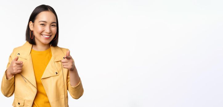 Hey you. Happy smiling asian woman pointing fingers at camera, choosing, congratulating and praising you, standing against white background.