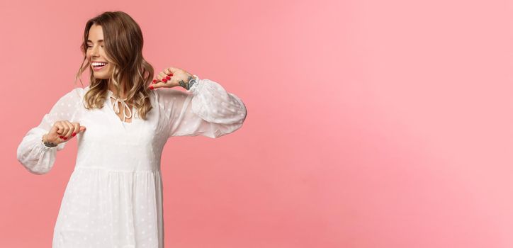 Beauty, fashion and women concept. Tender and carefree pretty young girl enjoying spring time, wearing white dress, dancing and looking away with beaming smile having fun, pink background.