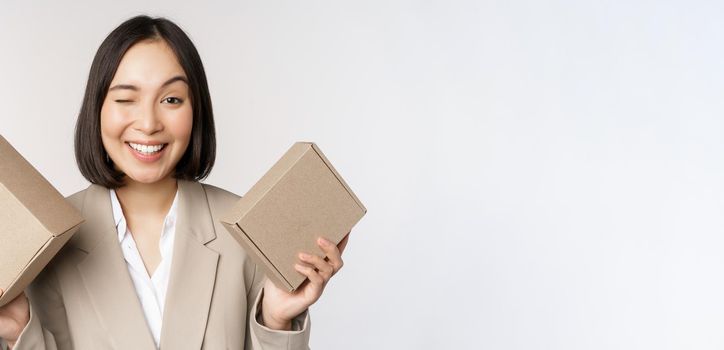 Image of saleswoman, asian businesswoman holding boxes with company brand product, smiling at camera, standing against white background.