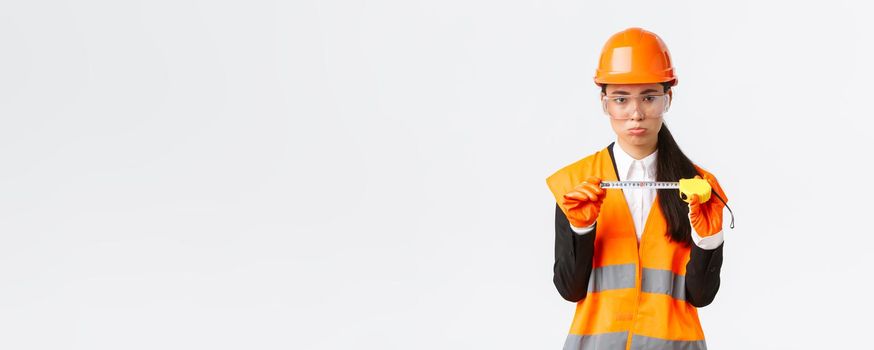 Disappointed gloomy female asian construction engineer, architect showing small size on tape measure, pouting upset with measurements, standing displeased in safety helmet, white background.