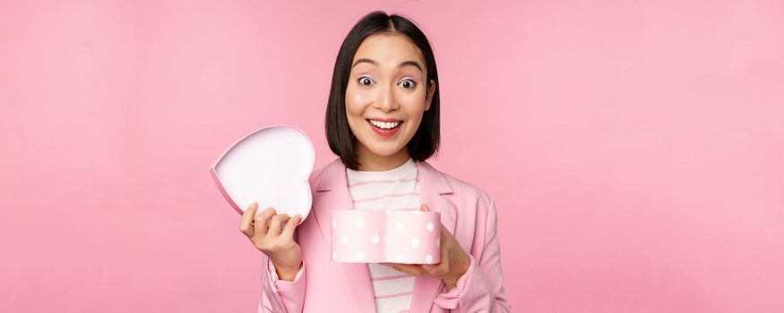 Happy cute korean girl in suit, opens up heart shaped box with romantic gift on white day holiday, standing in suit over pink background.