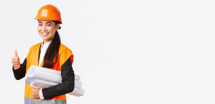 Confident successful female asian construction manager, architect in safety helmet and jacket, showing thumb-up and carry blueprints of building project, guarantee quality, white background.