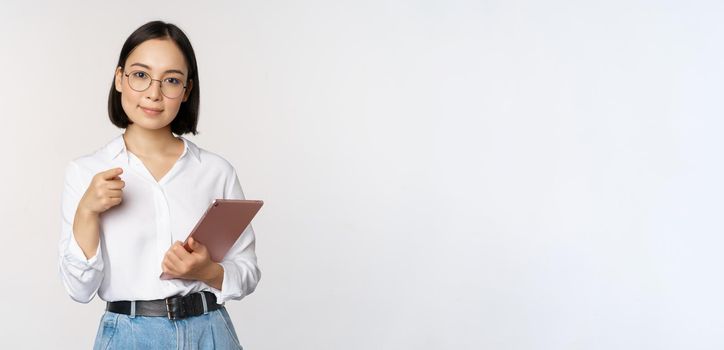 Korean woman, office worker manager in glasses, holding working tablet and pointing at you, choosing recruiting, standing over white background.