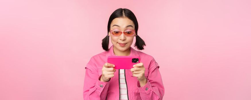 Portrait of happy asian girl playing on smartphone, watching videos on mobile phone app, standing over pink background.